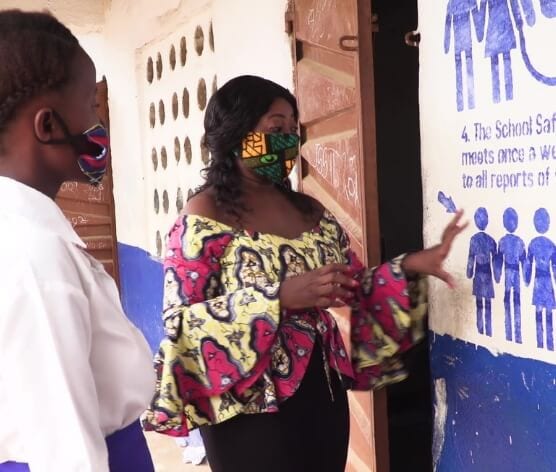 A teacher wearing a colourful mask is showing the school’s code of conduct painted in blue in a mural to a girl student.