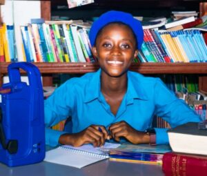 A smiling girl is looking at the camera while sitting at a desk and handwriting in her notebook. She’s wearing a blue uniform and a hat. There are several books on a shelf behind her and a radio on the desk.