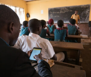 A classroom with a teacher doing a presentation to his pupils. In the first rows, some pupils dressed in dark green uniforms are paying attention. At the back, a supervisor is using a tablet to record monitoring data. 