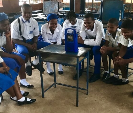 A group of girls and boys, in blue and white uniforms, are sitting in a circle in a classroom around a radio on top of a table. They are listening to the Girls and Boys Clubs programme.